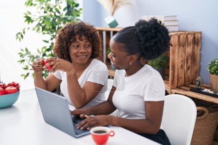 Foto de African american women mother and daughter drinking coffee using laptop at home - Imagen libre de derechos
