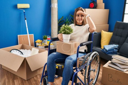 Foto de Young woman sitting on wheelchair moving to a new home very happy and smiling looking far away with hand over head. searching concept. - Imagen libre de derechos