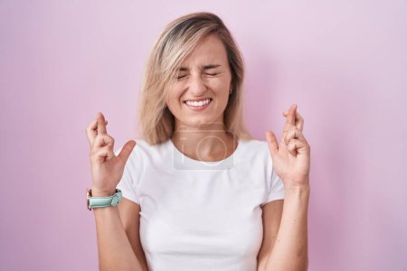 Foto de Young blonde woman standing over pink background gesturing finger crossed smiling with hope and eyes closed. luck and superstitious concept. - Imagen libre de derechos