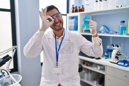 Téléchargez les photos : Jeune homme hispanique avec barbe travaillant au laboratoire scientifique tenant un ruban bleu souriant heureux faisant signe ok avec la main sur les yeux regardant à travers les doigts - en image libre de droit