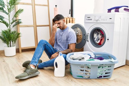 Foto de Young hispanic man putting dirty laundry into washing machine tired rubbing nose and eyes feeling fatigue and headache. stress and frustration concept. - Imagen libre de derechos