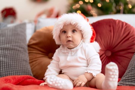 Foto de Adorable niño pequeño con sombrero de Navidad sentado en el sofá en casa - Imagen libre de derechos