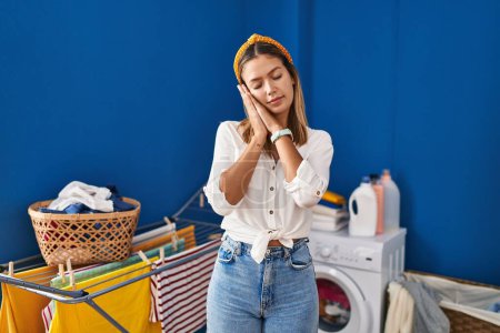 Téléchargez les photos : Young blonde woman at laundry room sleeping tired dreaming and posing with hands together while smiling with closed eyes. - en image libre de droit