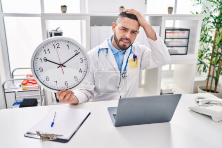 Foto de Hispanic doctor man holding clock at the clinic stressed and frustrated with hand on head, surprised and angry face - Imagen libre de derechos