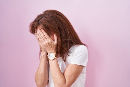 Foto de Mujer de mediana edad de pie sobre fondo rosa con expresión triste cubriendo la cara con las manos mientras llora. concepto de depresión. - Imagen libre de derechos