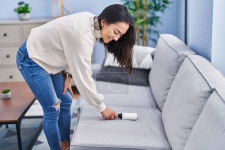 Photo for Young hispanic woman cleaning sofa using hair pet roller at home - Royalty Free Image