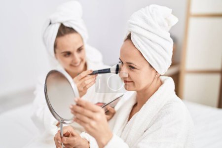 Photo for Two women mother and daughter wearing bathrobe applying skin makeup at bedroom - Royalty Free Image