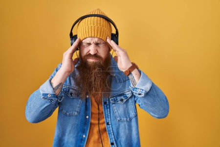 Foto de Caucasian man with long beard listening to music using headphones with hand on head, headache because stress. suffering migraine. - Imagen libre de derechos