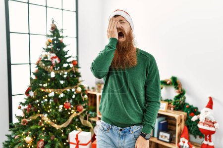 Foto de Redhead man with long beard wearing christmas hat by christmas tree yawning tired covering half face, eye and mouth with hand. face hurts in pain. - Imagen libre de derechos