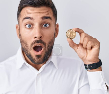 Foto de Handsome hispanic man holding litecoin cryptocurrency coin scared and amazed with open mouth for surprise, disbelief face - Imagen libre de derechos