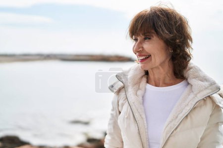 Téléchargez les photos : Femme d'âge moyen souriant confiant regardant vers le côté au bord de la mer - en image libre de droit