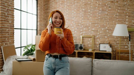 Photo for Young redhead woman talking on the smartphone drinking coffee at new home - Royalty Free Image