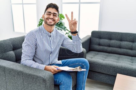 Photo for Young psychologist man at consultation office smiling with happy face winking at the camera doing victory sign with fingers. number two. - Royalty Free Image