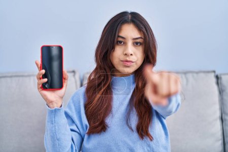 Téléchargez les photos : Jeune femme hispanique tenant smartphone montrant écran vide pointant du doigt vers la caméra et à vous, geste confiant regardant sérieux - en image libre de droit