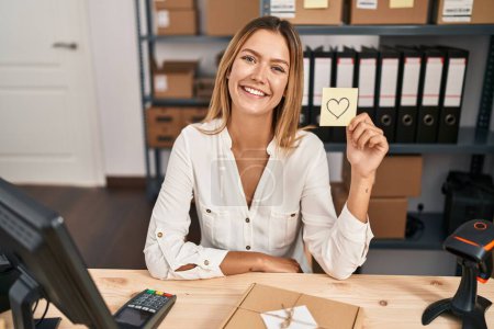 Téléchargez les photos : Young blonde woman working at small business ecommerce holding heart note looking positive and happy standing and smiling with a confident smile showing teeth - en image libre de droit
