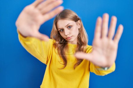 Foto de Joven mujer caucásica de pie sobre fondo azul haciendo marco usando las manos palmas y los dedos, perspectiva de la cámara - Imagen libre de derechos
