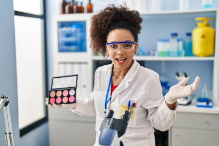 Photo for Young african american woman working at scientist laboratory doing make up celebrating victory with happy smile and winner expression with raised hands - Royalty Free Image