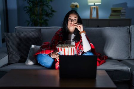 Téléchargez les photos : Femme hispanique manger du pop-corn regarder un film sur le canapé bouche et les lèvres fermées comme zip avec les doigts. secret et silencieux, tabou parlant - en image libre de droit