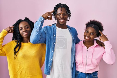 Foto de Grupo de tres jóvenes negros de pie juntos sobre fondo rosa sonriendo apuntando a la cabeza con un dedo, gran idea o pensamiento, buena memoria - Imagen libre de derechos