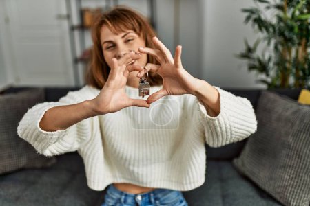 Téléchargez les photos : Jeune femme caucasienne souriante confiante tenant la clé de la nouvelle maison faisant symbole de coeur avec les mains à la maison - en image libre de droit