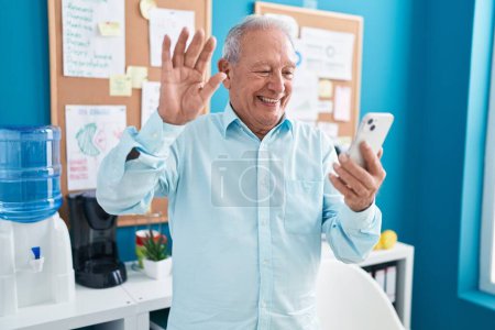 Foto de Senior man with grey hair working at the office doing video call with smartphone looking positive and happy standing and smiling with a confident smile showing teeth - Imagen libre de derechos