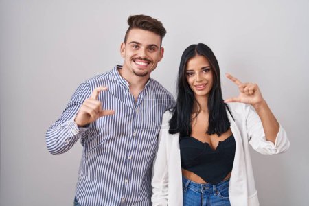 Foto de Young hispanic couple standing over white background smiling and confident gesturing with hand doing small size sign with fingers looking and the camera. measure concept. - Imagen libre de derechos
