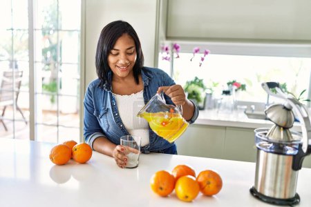 Foto de Mujer morena hispana bebiendo un vaso de jugo de naranja fresco en la cocina - Imagen libre de derechos
