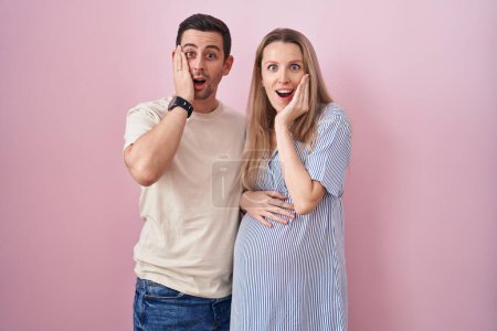 Photo for Young couple expecting a baby standing over pink background afraid and shocked, surprise and amazed expression with hands on face - Royalty Free Image