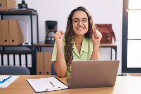 Téléchargez les photos : Young hispanic woman working at the office wearing glasses excited for success with arms raised and eyes closed celebrating victory smiling. winner concept. - en image libre de droit