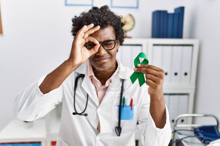 Foto de Africano médico hombre sosteniendo apoyo verde cinta sonriendo feliz haciendo ok signo con la mano en el ojo mirando a través de los dedos - Imagen libre de derechos
