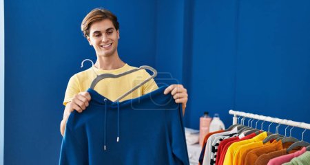 Photo for Young caucasian man smiling confident putting sweatshirt on hanger at laundry room - Royalty Free Image