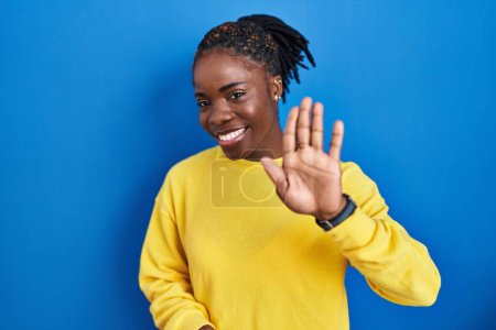 Téléchargez les photos : Belle femme noire debout sur fond bleu renonçant à dire bonjour heureux et souriant, geste de bienvenue amical - en image libre de droit