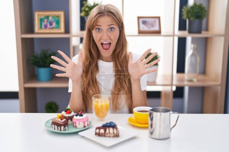 Téléchargez les photos : Jeune femme caucasienne mangeant des pâtisseries t pour le petit déjeuner célébrant fou et étonné pour le succès avec les bras levés et les yeux ouverts hurlant excité. concept gagnant - en image libre de droit