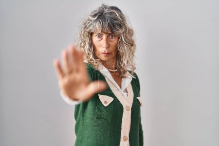 Foto de Middle age woman standing over white background doing stop sing with palm of the hand. warning expression with negative and serious gesture on the face. - Imagen libre de derechos