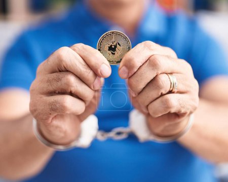 Photo for Middle age grey-haired man criminal holding uniswap crypto currency wearing handcuffs at home - Royalty Free Image