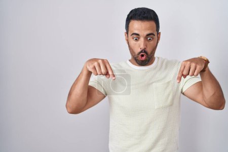 Photo for Hispanic man with beard standing over isolated background pointing down with fingers showing advertisement, surprised face and open mouth - Royalty Free Image