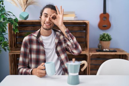Foto de Joven hombre hispano bebiendo café de la cafetera francesa sonriendo feliz haciendo signo bien con la mano en el ojo mirando a través de los dedos - Imagen libre de derechos