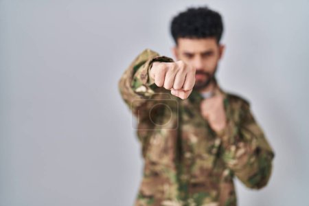Foto de Arab man wearing camouflage army uniform punching fist to fight, aggressive and angry attack, threat and violence - Imagen libre de derechos