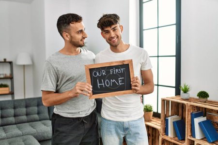 Photo for Two hispanic men couple hugging each other holding blackboard with our first home message at home - Royalty Free Image