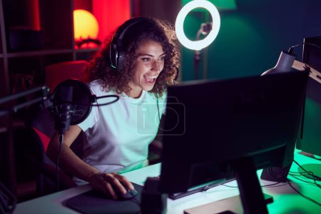Foto de Young beautiful hispanic woman streamer playing video game using computer at gaming room - Imagen libre de derechos