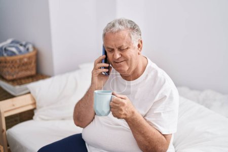 Téléchargez les photos : Moyen Age homme aux cheveux gris parler sur smartphone boire du café dans la chambre - en image libre de droit