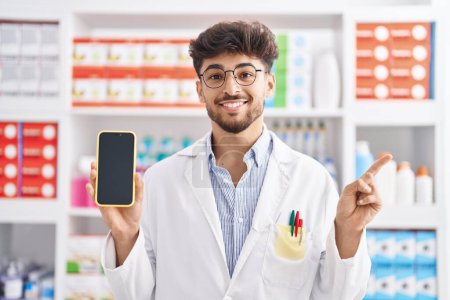Téléchargez les photos : Arabe homme avec barbe travaillant à la pharmacie pharmacie montrant écran smartphone souriant heureux pointant avec la main et le doigt sur le côté - en image libre de droit