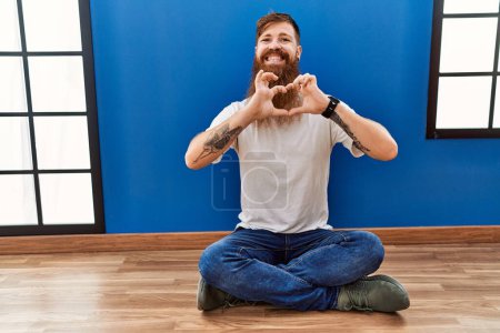 Foto de Redhead man with long beard sitting on the floor at empty room smiling in love doing heart symbol shape with hands. romantic concept. - Imagen libre de derechos