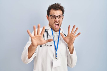 Photo for Young hispanic man wearing doctor uniform and stethoscope afraid and terrified with fear expression stop gesture with hands, shouting in shock. panic concept. - Royalty Free Image