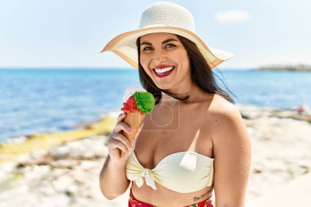 Foto de Joven mujer hispana hermosa toruist sonriendo confiado comiendo helado en la playa - Imagen libre de derechos