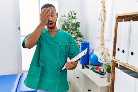 Foto de African american physiotherapist man working at pain recovery clinic yawning tired covering half face, eye and mouth with hand. face hurts in pain. - Imagen libre de derechos