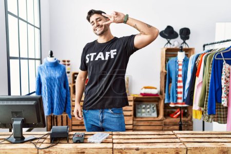 Foto de Joven hombre hispano que trabaja en una boutique minorista haciendo símbolo de paz con los dedos sobre la cara, sonriendo alegre mostrando la victoria - Imagen libre de derechos