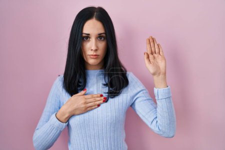 Foto de Hispanic woman standing over pink background swearing with hand on chest and open palm, making a loyalty promise oath - Imagen libre de derechos