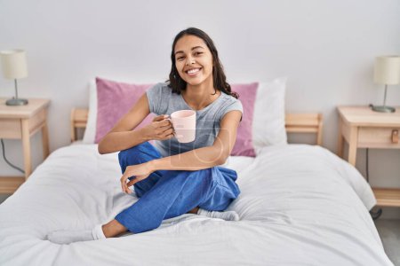 Téléchargez les photos : Young african american woman drinking cup of coffee sitting on bed at bedroom - en image libre de droit