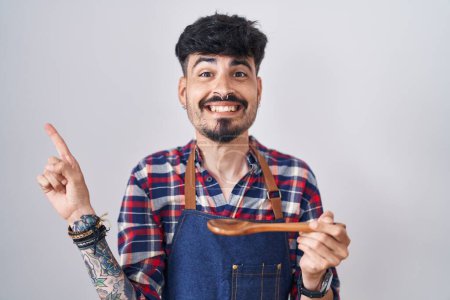 Photo for Young hispanic man with beard wearing apron tasting food holding wooden spoon smiling happy pointing with hand and finger to the side - Royalty Free Image